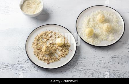 Weiße Schokoladentrüffel in Kokosflocken gerollt und zerdrückte Nüsse auf einem weißen Teller. Kochstufe mit köstlichen Pralinen Stockfoto