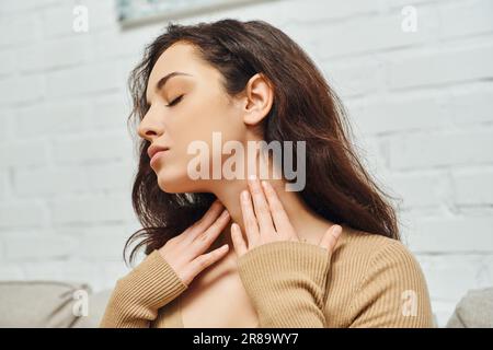 Junge brünette Frau in lässiger Kleidung, die den Hals berührt, während sie die Schilddrüse überprüft und zu Hause auf der Couch sitzt, konzentriert sich auf Selbstpflege und Stockfoto