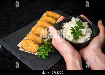 Kabeljauknödel oder „Bolinhos de bacalhau“, sehr berühmt in der portugiesischen Gastronomie. Hände halten Topf mit weißem Reis. Stockfoto