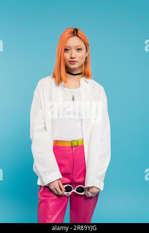 Junges asiatisches Modemodell in weißem Hemd, pinkfarbenen Hosen und trendigen Accessoires mit Sonnenbrille und Blick auf die Kamera auf blauem Hintergrund, trendiger Sommer Stockfoto