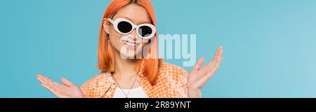 Positive, junge asiatische Frau mit gefärbtem Haar, in lässiger Kleidung und Sonnenbrille, Gesten mit Händen auf leuchtend blauem Hintergrund, orangefarbenes Hemd, ne Stockfoto