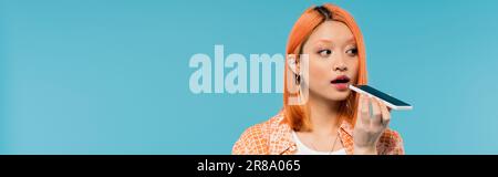 Social-Media-Influencer, junge asiatische Frau mit gefärbtem Haar, hält Smartphone und sendet Sprachnachricht auf blauem Hintergrund, Mobiltelefon, Jugendkultur Stockfoto