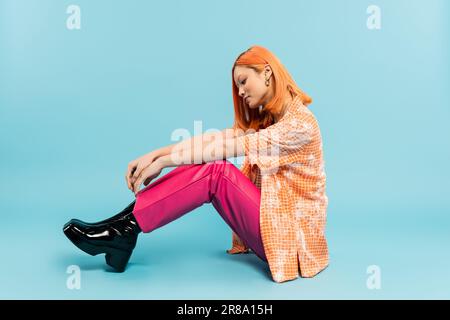 Seitenansicht einer jungen und stilvollen asiatischen Frau in orangefarbenem Hemd und pinkfarbener Hose auf blauem Hintergrund, gefärbtes rotes Haar, Sommermode, jugendlicher Stil Stockfoto