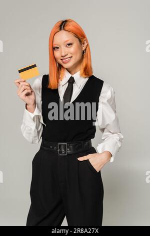 Fröhliche Emotionen, junge und fröhliche asiatische Frau, die mit der Hand in der Tasche steht und Kreditkarte auf grauem Hintergrund zeigt, schwarze und weiße Kleidung, Busin Stockfoto