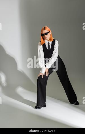 Stilvolle Pose einer modischen asiatischen Frau mit rotem Haar, dunkler Sonnenbrille, weißem Hemd, schwarzer Krawatte, Weste und Hose auf grauem Hintergrund, b Stockfoto