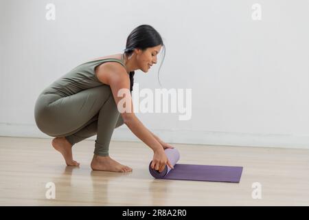 Schlanke Frau lächelt, während sie ihre Matte nach dem Yoga-Training weglegt Stockfoto