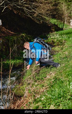 Wanderer trinken Flusswasser in den Bergen. Ein erwachsener Mann trinkt Wasser aus dem Bergbach. Touristen, die lässige Kleidung und Sonnenbrille tragen und einen Schluck trinken Stockfoto