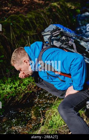 Wanderer trinken Flusswasser in den Bergen. Ein erwachsener Mann trinkt Wasser aus dem Bergbach. Touristen, die lässige Kleidung und Sonnenbrille tragen und einen Schluck trinken Stockfoto