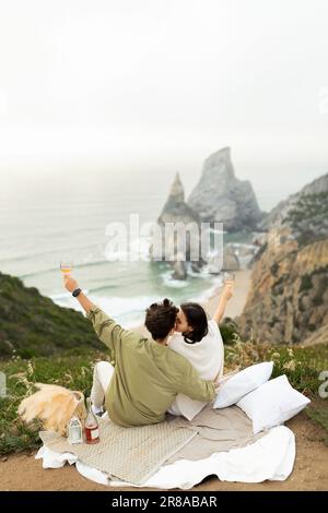 Ein Paar, das seine Verlobung an der Küste feiert, Champagner trinkt und einen atemberaubenden Blick auf die Küste hat Stockfoto