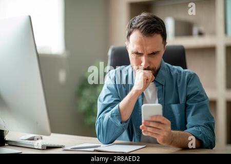 Gestresster Geschäftsmann, der auf den Smartphone-Bildschirm schaut, während er im Büro am Schreibtisch sitzt Stockfoto
