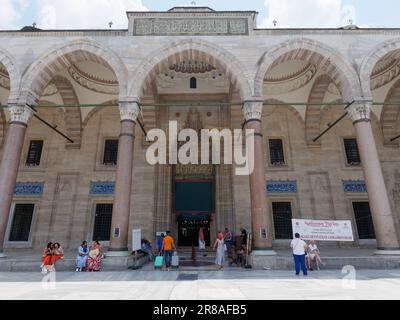 Touristen am Haupteingang der Suleymaniye Moschee in Istanbul, Türkei. Ein Mann bringt kleine Koffer zur Moschee, während die Leute draußen sitzen Stockfoto