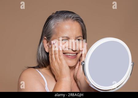 Eine lächelnde europäische ältere grauhaarige Frau genießt natürliche Hautschönheit und Lebensstil. Schauen Sie in den Spiegel Stockfoto