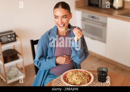 Erwartungsvolle Freuden. Glückliche, junge schwangere Frau, die hausgemachte Pasta isst, am Tisch in der Küche sitzt Stockfoto