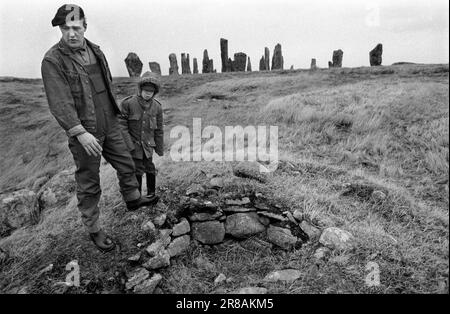 Callanish Standing Stones, Isle of Lewis, Outer Hebrides Schottland, ca. Juni 1974. Die Callanish Stones sind eine Anordnung von Steinen in einem Kreuzmuster mit einem zentralen Steinkreis. Sie wurden in der späten Jungsteinzeit errichtet und waren ein Schwerpunkt für rituelle Aktivitäten während der Bronzezeit. Vater und Sohn der Dorfbewohner weisen auf einen liegengebliebenen Begräbniszist hin - einen kleinen, aus Stein gebauten Sarg. 1970S GB HOMER SYKES Stockfoto