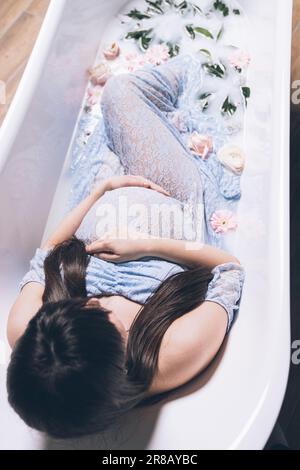 Eine junge, schwangere Frau sitzt in einer luxuriösen Badewanne, umgeben von farbenfrohen Rosenblättern, und genießt ein beruhigendes, aromatisches Erlebnis Stockfoto