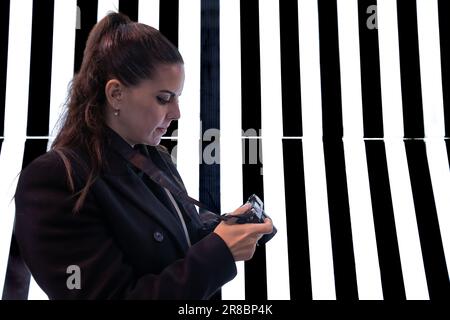 Eine junge brünette Frau, die die Fotos auf ihrer Kamera vor einem leuchtenden Hintergrund mit schwarzen und weißen vertikalen Streifen betrachtet. Fotografiekonzept Stockfoto