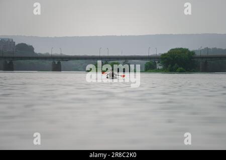 Junger Mann paddelt in einem grünen Kajak mit orangefarbenen Paddeln in einem Fluss mit Bergen, Bäumen und einer Brücke in der Ferne Stockfoto