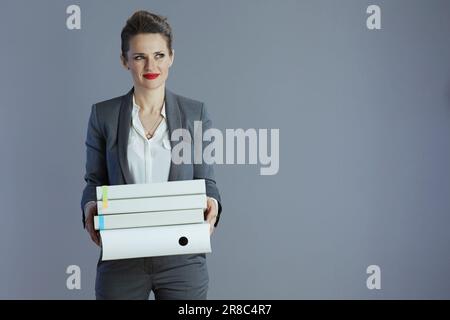 Nachdenkliche, trendige Geschäftsfrau mittleren Alters im grauen Anzug mit isolierten Ordnern auf grauem Hintergrund. Stockfoto