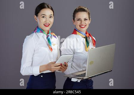 Lächelnde moderne Stewardess in blauem Rock, weißem Hemd und Schal mit Tablet-PC und Laptop isoliert auf grauem Hintergrund. Stockfoto