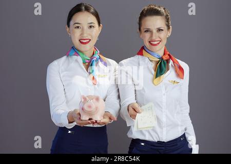 Lächelnde, stilvolle Stewardess in blauem Rock, weißem Hemd und Schal mit Flugtickets und Schweinebank isoliert auf grauem Hintergrund. Stockfoto