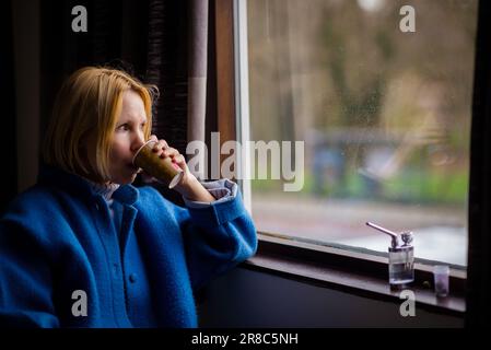 Eine Frau trinkt Kaffee und raucht Cannabis. Eine Erwachsene Frau über 40 sitzt neben dem Fenster, daneben befindet sich ein Gerät zum Rauchen von Marihuana. Stockfoto