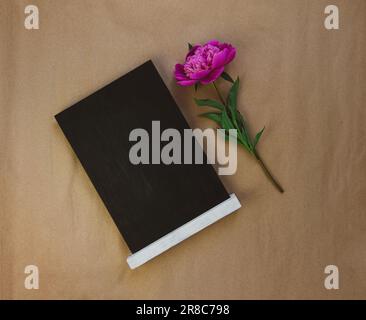 Leere Tafel mit pinkfarbener Pfingstrose auf braunem Kraftpapierhintergrund. Tafel mit wunderschöner Blumentapete zum Kopieren. Minimalistischer Style. Birt Stockfoto