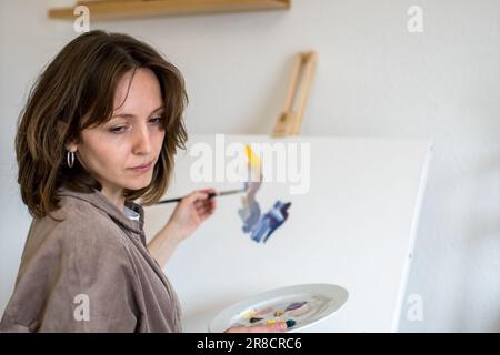 Bild einer Malerin, die ihre Notizen ansieht, während sie zu Hause einen Pinsel und eine Farbpalette mit Acryl auf einer weißen Leinwand hält Stockfoto