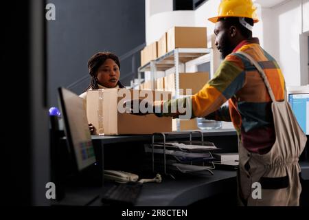 Postdienstmitarbeiter, der den Empfang und die Registrierung von Paketen im Lager verwaltet. Eine afroamerikanische Kurierin stellt Pappkartons auf den Tresen im Lager der Versandfirma Stockfoto