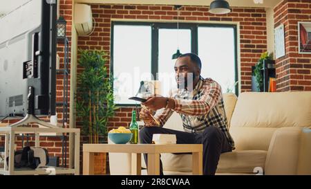 Ein afroamerikaner isst Nudeln im Wohnzimmer, sitzt im Fernsehen und sieht sich Filme an. Junger Mann, der asiatische Speisen mit Essstäbchen serviert, bestellt Abendessen aus dem Restaurant zum Mitnehmen. Stockfoto