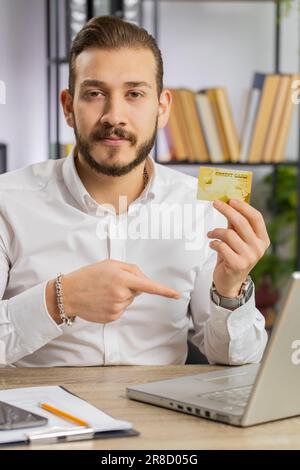 Ein Geschäftsmann zeigt Kreditkartenwerbung, die Geld bargeldlos auf einem Laptop überträgt, Einkäufe im Internet, Rabatte, Internetbestellungen. Ein Typ am Arbeitsplatz. Vertikal Stockfoto