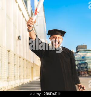 Glücklicher erwachsener Mann mit Abschluss, der ein Junggesellenkleid und ein schwarzes Mörtelbrett trägt und sein Diplom zeigt, während er in die Kamera schaut Stockfoto