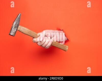 Die Hand eines Mannes hält einen Hammer durch zerrissenes orangefarbenes Papier. Konzept, Hand durch zerrissenes orangefarbenes Papier. Stockfoto