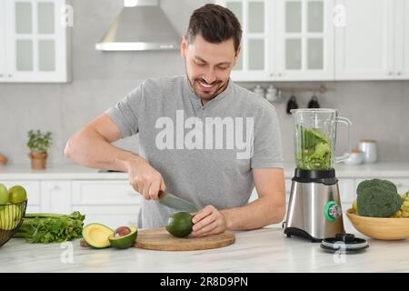 Ein glücklicher Mann schneidet Avocado für köstlichen Smoothie an einem weißen Marmortisch in der Küche Stockfoto