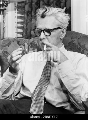 New York, New York: 1954 bekannter Autor, Karikaturist und Humorist James Thurber sitzt auf einem Stuhl und zündet eine Zigarette an. Stockfoto