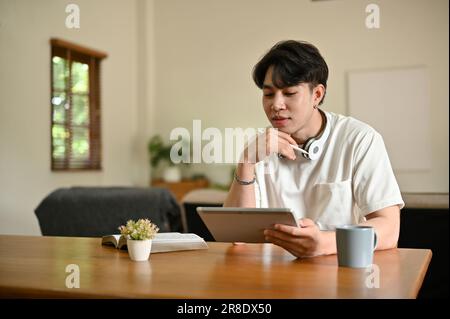 Ein gutaussehender und glücklicher junger asiatischer Student oder Freiberufler, der sein digitales Tablet an einem Tisch in seinem Wohnzimmer verwendet. Stockfoto