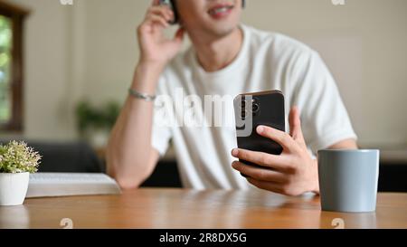 Nahaufnahme eines Asiaten in legerer Kleidung, der sein Smartphone hält und Musik über Kopfhörer an einem Tisch in seinem Zimmer hört. Stockfoto