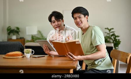 Ein Porträt von zwei jungen asiatischen Männern in legerer Kleidung, die in die Kamera schauen und mit einem digitalen Tablet und einem Buch in der Hand an einem Tisch sitzen. Stockfoto