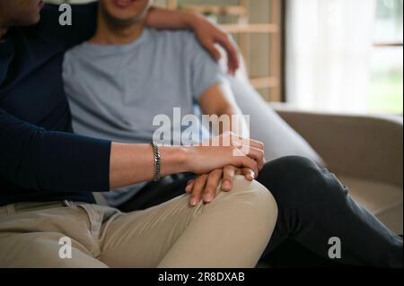 Nahaufnahme eines schwulen Mannes, der die Hand seines Freundes hält, während er sich entspannt und einen Film auf einer Couch im Wohnzimmer ansieht. LGBTQ+, Beziehungen Stockfoto