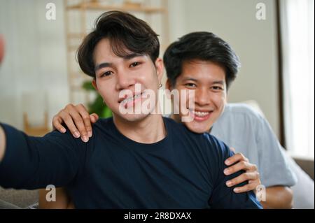 Ein Porträt eines reizenden und glücklichen, jungen asiatischen, schwulen Paares zu Hause, das sich umarmt und glücklich in die Kamera lächelt. LGBTQ+, glücklicher Moment, sorgenfrei Stockfoto