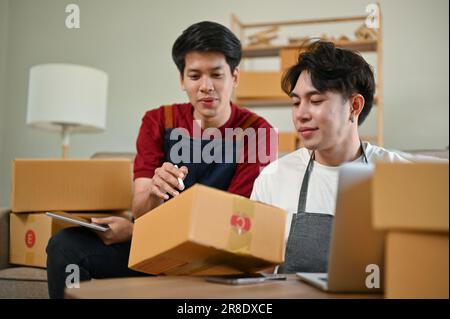 Ein glücklicher junger asiatischer Mann, der Versandbestellungen überprüft und vorbereitet, Versandkartons verpackt und mit seinem Freund zu Hause arbeitet. Unternehmer, Busine Stockfoto