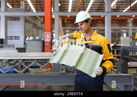 Junge hispanische Arbeiter in Polyester-Jacke Uniform, Schutzhelm und Schutzhandschuh prüfen die Qualität von Wellblechdächern. Das Metall Stockfoto