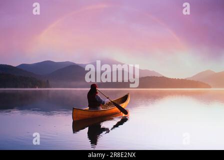 USA, St. Armand, Rückansicht einer Frau, die Kanu auf dem ruhigen Lake Placid bei Sonnenuntergang im Adirondack Park fährt Stockfoto