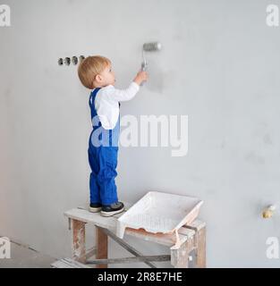 Kleiner Bauarbeiter, der die Wand in einer Wohnung streicht, die gerade renoviert wird. Ein niedliches Kind im Arbeitsanzug, das Malerwalze benutzt, während es zukünftig zu Hause spielt. Stockfoto