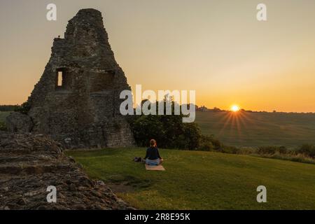 Hadleigh Castle, Großbritannien. Juni 2023. Ein ruhiger Sonnenaufgang am Hadleigh Castle in Essex mit einer einsamen Figur, die in der Nähe der antiken Ruinen meditiert. Das goldene Licht der Dämmerung wirft lange Schatten über den grasbewachsenen Hügel und ruft Themen der Reflexion, Erneuerung und Verbindung zum Ort hervor. Sommersonnenwende an den Ruinen der Burg in der Stadt. Penelope Barritt/Alamy Live News Stockfoto