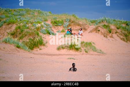 Menschen genießen das warme Wetter an einem anderen Ort, Anthony Gormleys Ironmen Statuen am Crosby Beach, Merseyside, am Vorabend der Sommersonnenwende. Foto: Dienstag, 20. Juni 2023. Stockfoto