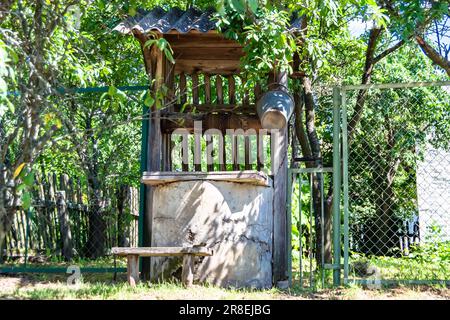 Alter Brunnen mit Eisenkübel auf langer geschmiedeter Kette für sauberes Trinkwasser, Fotografie bestehend aus altem abgerundetem Brunnen mit Dach, klarem Wasser in großem Bock Stockfoto