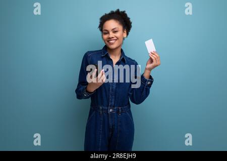 Junge, charmante, brünette lateinische Frau in blauem Jeansanzug mit Kreditkartenmodell und Smartphone Stockfoto
