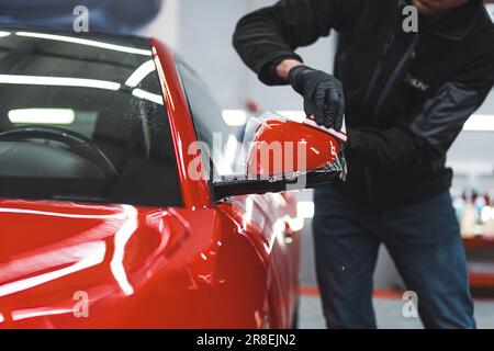 Spezialist für Autoverpackungen in Schutzhandschuhen, der den roten Außenspiegel des Fahrzeugs mit transparenter Folie umhüllt, um den Lack zu schützen. Hochwertiges Foto Stockfoto