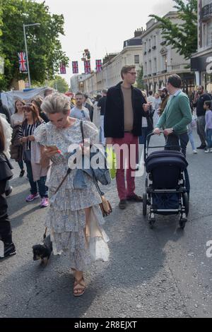 Frau, die einen Hund begleitet und gleichzeitig ein iphone liest. Die Kings Road, Platinum Jubilee Street Party, die den Aufruf zur Krise des britischen Roten Kreuzes in der Ukraine unterstützt. Die Kings Road ist zum Teil für den Verkehr gesperrt, lokale Chelsea und Knightsbridge junge Erwachsene unterhalten sich, während eine Frau mit ihrem Hund geht und liest oder iphone. Chelsea, London, England 4. Juni 2022. HOMER SYKES AUS DEN 2020ER JAHREN Stockfoto