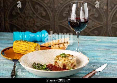 Argentinisches Entrana-Steak unter Käse, gegrilltes Rindfleisch mit Spargel, Mais und Tomatensouce auf hölzernem blauem Hintergrund Stockfoto